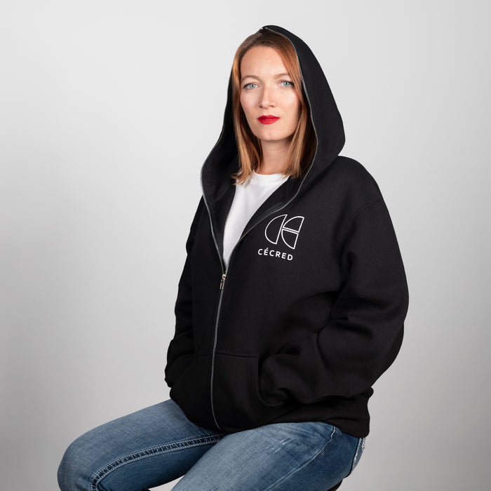 Woman seated wearing the Cécred black zip-up hoodie with hood up, styled casually with denim, chest logo visible