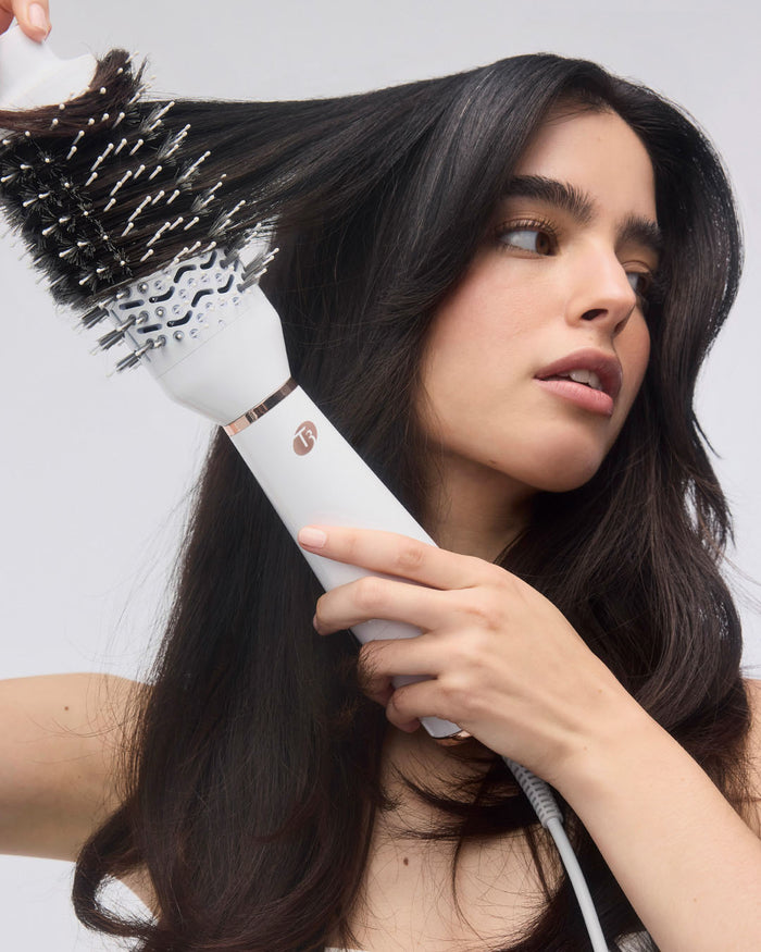 Woman using a white hair dryer brush on her hair against a plain background