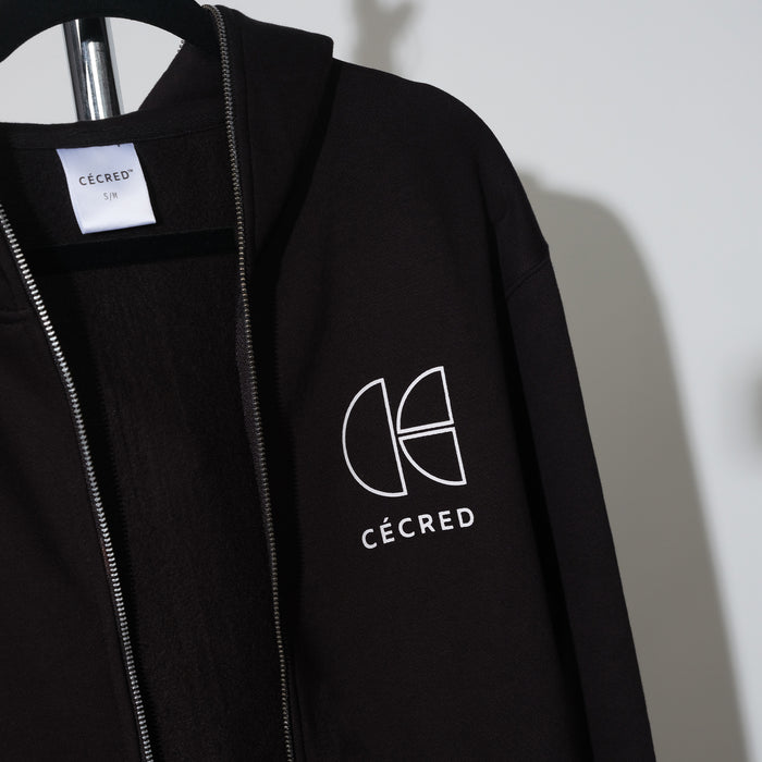 Close-up of the Cécred neck tag showing size label, with the collar and chest logo visible on the black zip-up hoodie