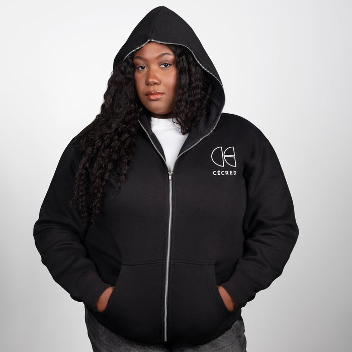 Woman modeling the Cécred black zip-up hoodie with hood up, front zipper closed, and chest logo visible