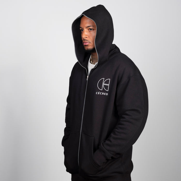 Man modeling the Cécred black zip-up hoodie with hood up, angled side view showing relaxed unisex fit and chest logo