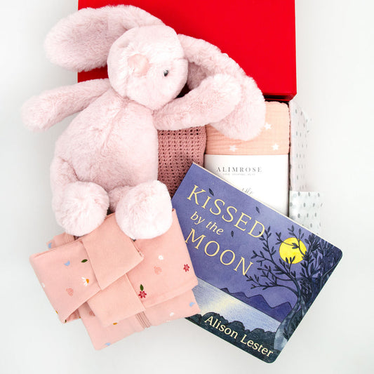 A collection of baby items including a pink plush rabbit, a pink starry night muslin swaddle, a pink cellular blanket, a pink and white romper, and a board book titled 'Kissed by the Moon'. All items are displayed in a red memory box. Price: 159