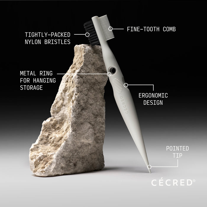 White comb with fine and coarse teeth, metal hanging ring, and pointed tip displayed against textured stone.