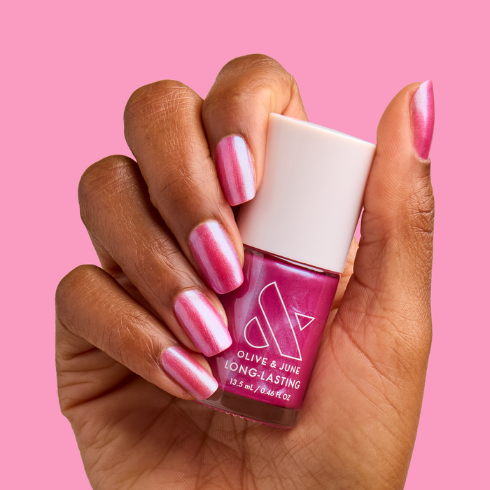 Hand with medium-length nails painted in HARD LAUNCH pink-purple iridescent duochrome polish holding bottle against pink background