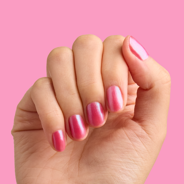 Hand with short nails painted in HARD LAUNCH pinky-purple iridescent duochrome polish against pink background