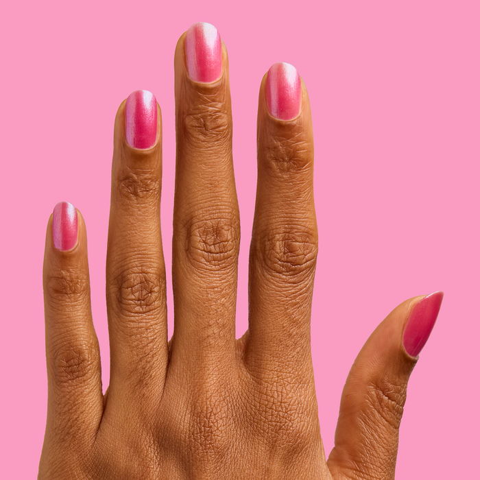 Hand with medium-length nails painted in HARD LAUNCH pinky-purple iridescent duochrome polish on a solid pink background