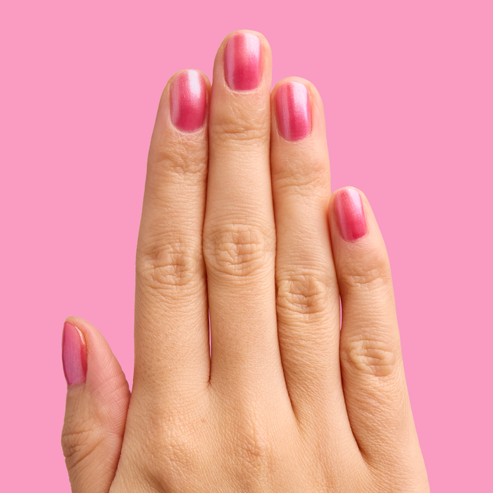 Hand with short nails painted in HARD LAUNCH pinky-purple iridescent duochrome polish on solid pink background