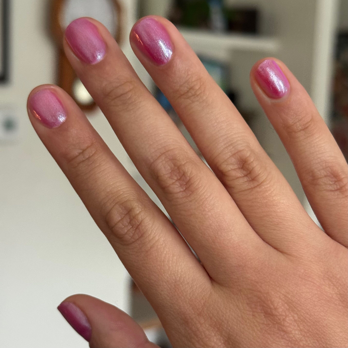 Hand with short nails painted in HARD LAUNCH pinky-purple iridescent duochrome polish on blurred indoor background