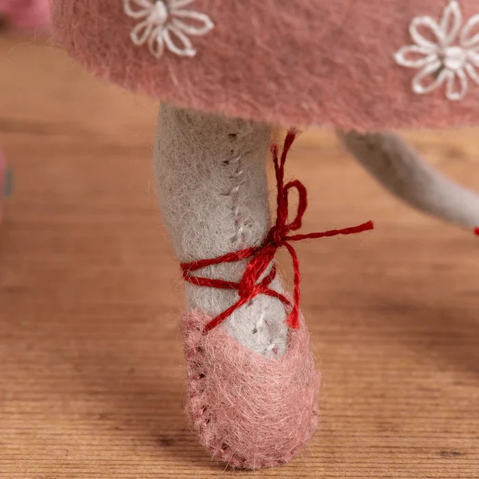 Close-up of a toy figure's foot wearing a pink ballet slipper with red ribbons on a wooden surface.