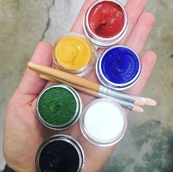 Hand holding six small containers of different colored face paint with applicators.