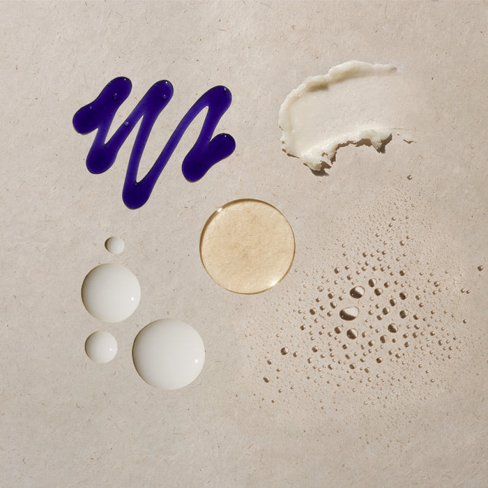Dark blue brushstroke, white cream swatches, beige circular swatch, white droplets, and tan textured spray on neutral backgro